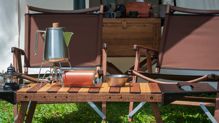 Stainless steel kettle, portable gas stove, bowl and vintage lanterns with outdoors wooden table set in front of tent on green lawn in camping area