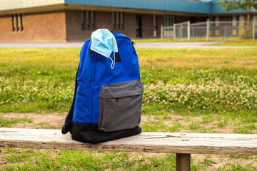 School backpack with medical mask on the old wooden bench. Elementary school in the background. Back to school during epidemic concept.
