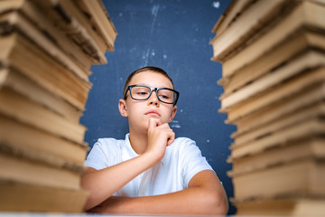 Smart boy in glasses sitting between two piles of books and look down thoughtfully.