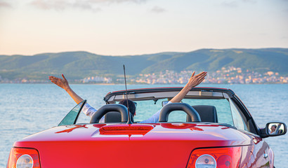 A woman in a red car raised her hands up at the seaside.