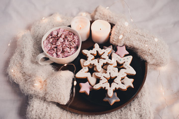 Cup of coffee with marshmallow, burning candles and ginger breads in star shape on wooden tray on knitted sweater closeup. Winter holiday season. .