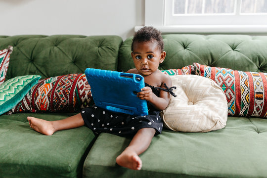 A little adorable toddler sitting on the couch, playing with a tablet.