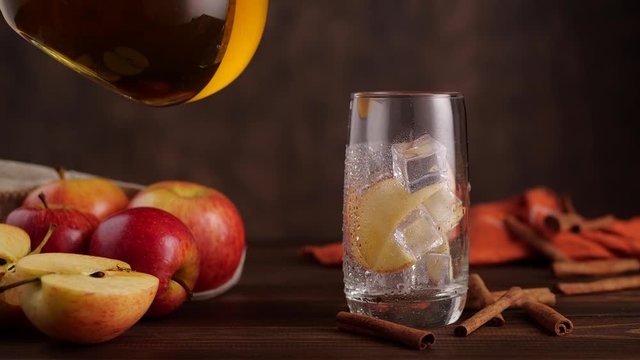 Unrecognizable person pouring fresh juice from jug into glass with ice near apples and cinnamon sticks