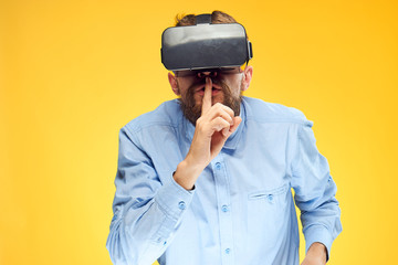 A man in virtual reality glasses in a blue shirt holds a finger near his mouth yellow background