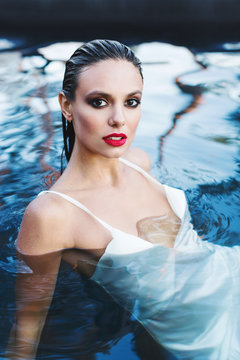 Beautiful brunette woman in white wet dress lies in the water. Woman in the pool