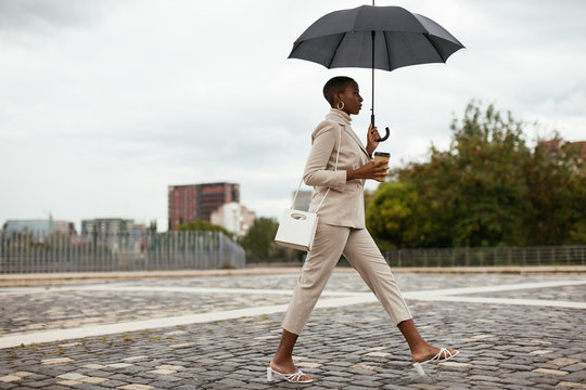 Confident black lady with umbrella and beverage walking on pavement