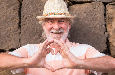 Attractive senior man white hair and beard smiling making heart shape with hands standing against a stone wall with straw hat - concept of active elderly during retirement