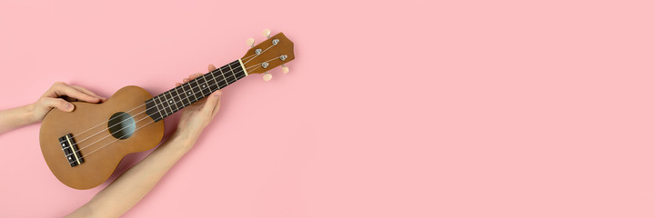 Female hands hold ukulele on a pastel pink background. Creative banner with copy space.