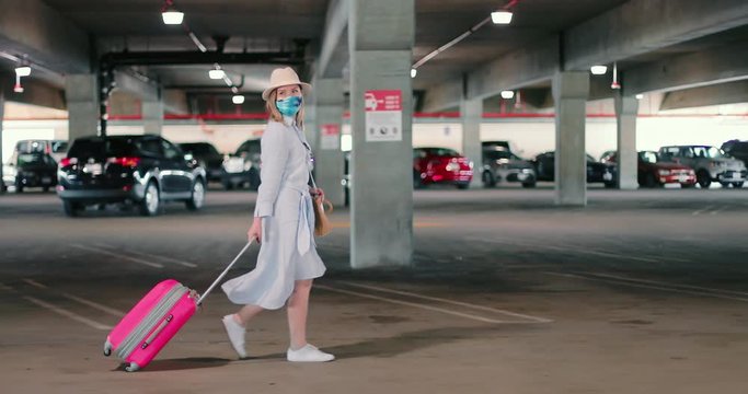 Safe travels under COVID-19. Slow motion of woman in face mask and trendy outfit walking by empty airport parking and carrying her bright pink suitcase on the wheels before departure to vacation
