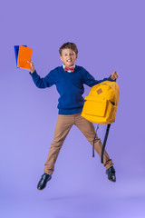 cheerful schoolboy in uniform with backpack and books jumps on purple background