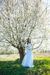 Beautiful girl in a light summer blue long dress adorned in her hair against a blossoming tree. Tender portrait of a young woman in white blossom