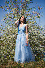 Fototapeta na wymiar Portrait of a tender girl in a blue long dress with tulle under blooming cherry with a smile on her face on a sunny warm summer day