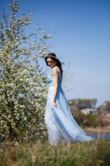 Fototapeta premium Portrait of a tender girl in a blue long dress with tulle under blooming cherry with a smile on her face on a sunny warm summer day