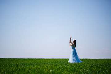 Fototapeta premium Woman in a blue long dress on a background of green field. Fashion portrait of a beautiful girl with a smile on her face