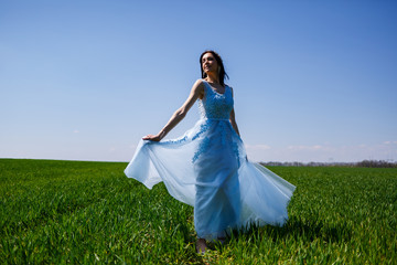 Fototapeta premium Young woman in a blue long dress on a background of green field. Fashion portrait of a beautiful girl with a smile on her face