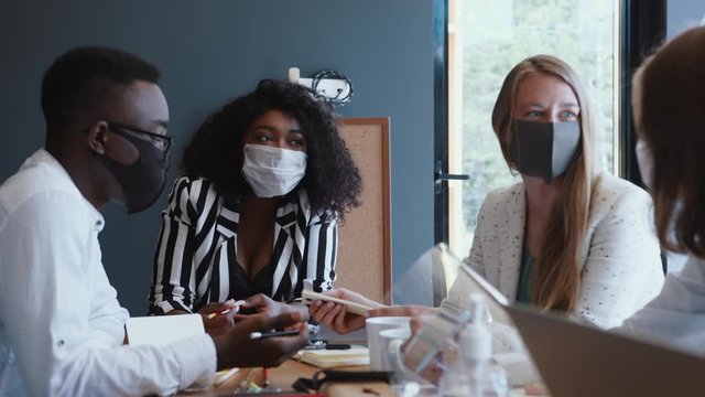 Happy multiethnic creative start-up business people talk at meeting at light modern office table wearing face masks.