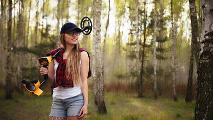 A young woman with a smile holding a metal detector over her shoulder in the forest. High quality...