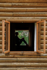 Silhouette of a young woman in the window of a wooden house in the mountains. Calm rest, alone with nature. In harmony with yourself. Meditation. Solitude in the mountains.