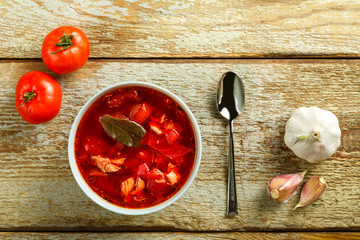 A plate of borscht with chicken with a spoon on a wooden table next to garlic and tomatoes.