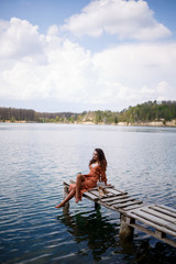 Fototapeta na wymiar A girl with long wavy curly hair in a long guipure dress barefoot in the summer in a forest on a lake at sunset standing on a pantone on a wooden pier bridge. Summer sunny day
