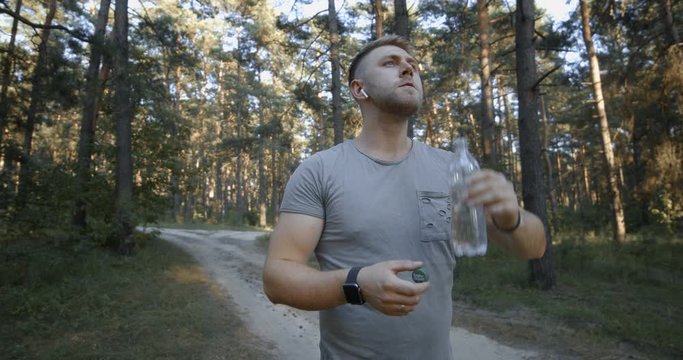 Young bearded man in sport outfit drinking water while standing at green forest. Muscular guy refreshing after regular workout on fresh air.