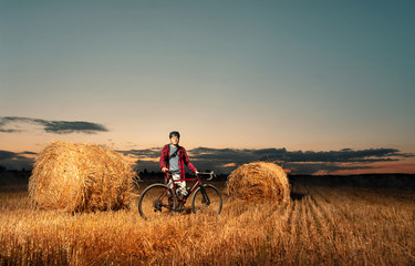 Obraz premium A cyclist in a helmet with bicycle stands in a field with bales of straw at sunset. Active and sporty lifestyle concept.