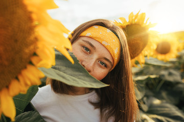 Attractive mysterious young woman look straight on camera covering her mouth with green leaf. Stand alone among sunflowers growing up on field. Harvest time in July or August.