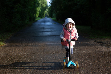Pretty toddler girl wearing rubber boots, grey hoodie and pink waistcoat riding scooter in the...