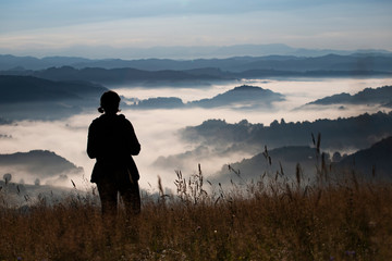 Young woman standing on golden meadow and watching towards the sunrise above mist.