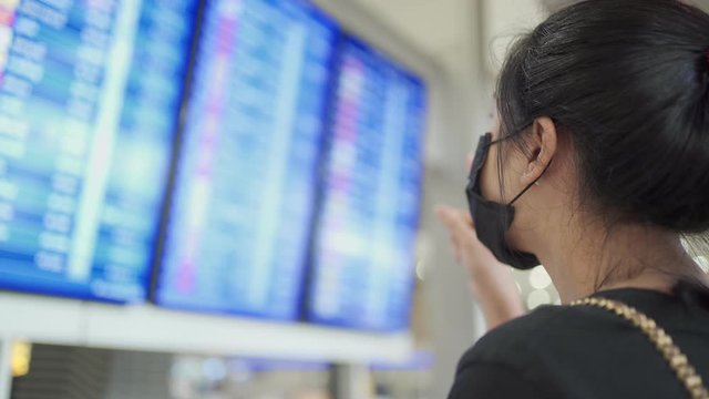 Asian woman wear medical mask standing in front of airlines flight time schedule screen at airport departure terminal, risk of public transpo corona virus covid19 pandemic, new normal social distance