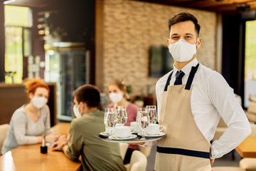 Happy waiter serving coffee to his customers while wearing protective face mask in a cafe