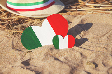 two wooden hearts painted in colors of the flag of Italy green, white and red and straw hat with ribbon stands on golden sand with straw on on Mediterranean coast, tourism concept, national day