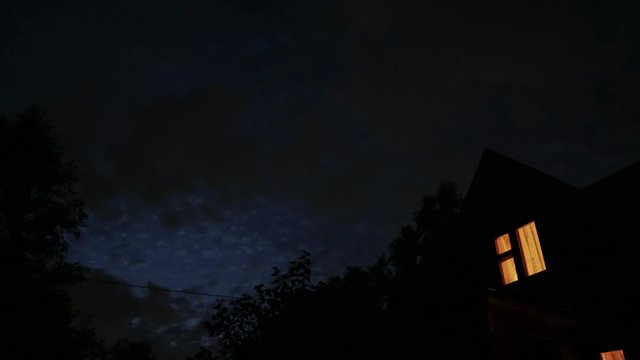 louds moving across the evening sky. Silhouette of a house, a window is lit in the house