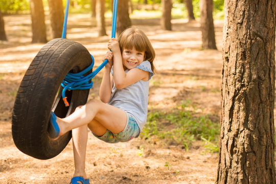 Happy active child girl playing on swing wheel in forest on sunny summer day. Preschool child having fun and swinging on a tire
