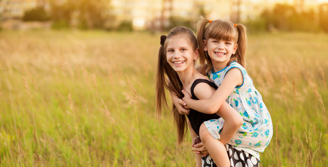 Fototapeta premium Happy child girl holding little sister in arms on back. Happy childhood together in summer time