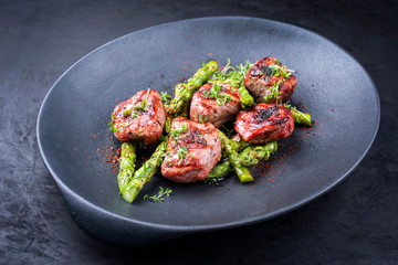 Traditional barbecue Iberian pork filet medaillons with green asparagus tips offered as close-up on a modern design cast iron black plate