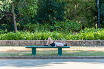 Mulher deitada sobre banco de concreto em parque da cidade de São Paulo, relaxando ouvindo musica com fones de ouvido e celular. 