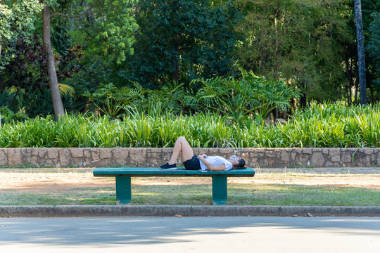 Mulher deitada sobre banco de concreto em parque da cidade de S&atilde;o Paulo, relaxando ouvindo musica com fones de ouvido e celular. 