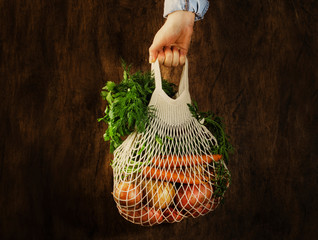 Mesh bag with vegetables and herbs in female hand. Woman hold string net shopping bag on wooden...