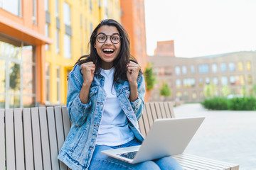 Portrait of excited joyful girl sitting on a bank outdoors with her laptop computer and celebrating...