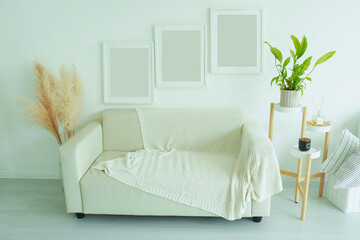 interior poster mock up with three vertical empty white frames, gray sofa with knitted blanket, houseplants and fragrance on stand in living room with white wall..