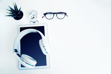 white wireless headphones on a white tablet, glasses, an alarm clock and a houseplant on a white...