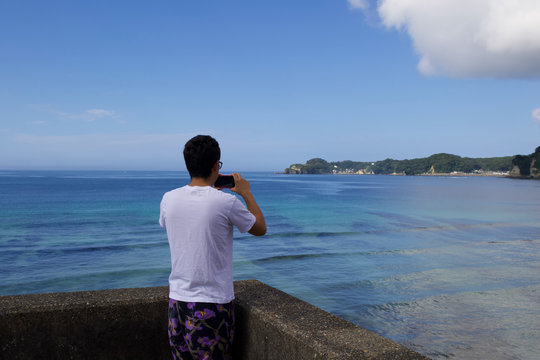 An Asian boy is taking pictures of a beautiful blue bay in Japan with his mobile device, it is a pretty scene with a wonderful landscape. Katsuura in Chiba is known for its many bays.