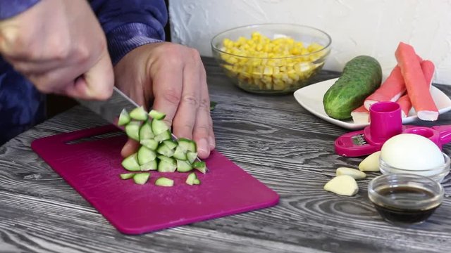 A man cuts a cucumber on a cutting board. Crab sticks, chicken eggs and vegetables for salad preparation. They lie on painted pine boards
