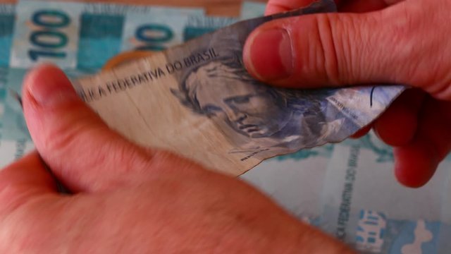 Brazilian currency, One hundred Real money lying on the desk and a battered two Reais banknote. The hand picks up the banknote and unfolds and smooths it