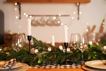 The festive Christmas table is decorated with branches of a Christmas tree, candles and garlands. Cozy home Christmas atmosphere.