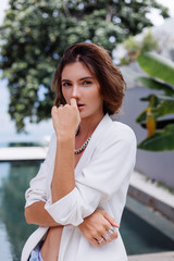 Fashion portrait of woman at tropical luxury villa wearing white stylish blazer, rings, earings on background of tropical leaves and swimming pool.