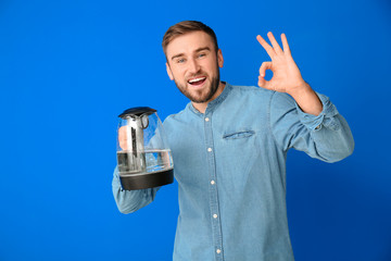 Young man with electric kettle showing OK gesture on color background