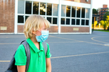 Kid in mask during corona virus outbreak. Little schoolboy wears grey backpack, breathes through mask, going to school. Back to school concept after reopening. Student at the school yard