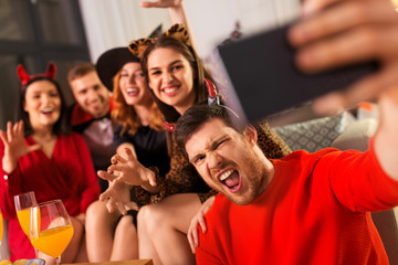 friendship, holiday and people concept - group of happy smiling friends in halloween costumes of vampire, devil, witch and cheetah taking selfie by smartphone at home party at night